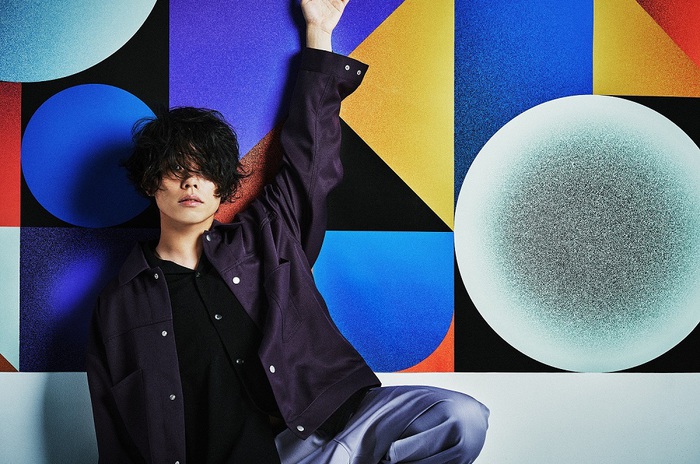 Musician Yoh Kamiyama sits in front of a colorful abstract wall with his left hand raised. His short black hair covers his eyes.