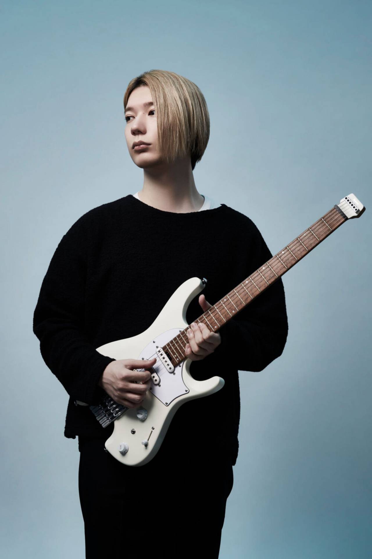 Ichika Nito wears all black holding his white signature headless guitar in front of a blue and white background.